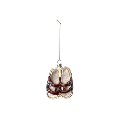 A glass ornament in the shape of a pair of sandals, with brown straps and gold-colored metal hardware, hanging from a gold-colored string.