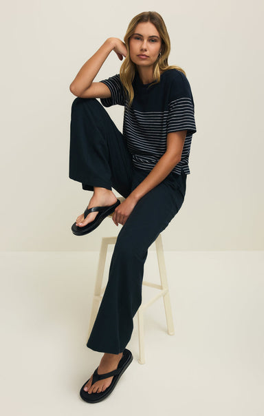 Woman sitting on a stool wearing a black and white striped shirt and black pants against a white background