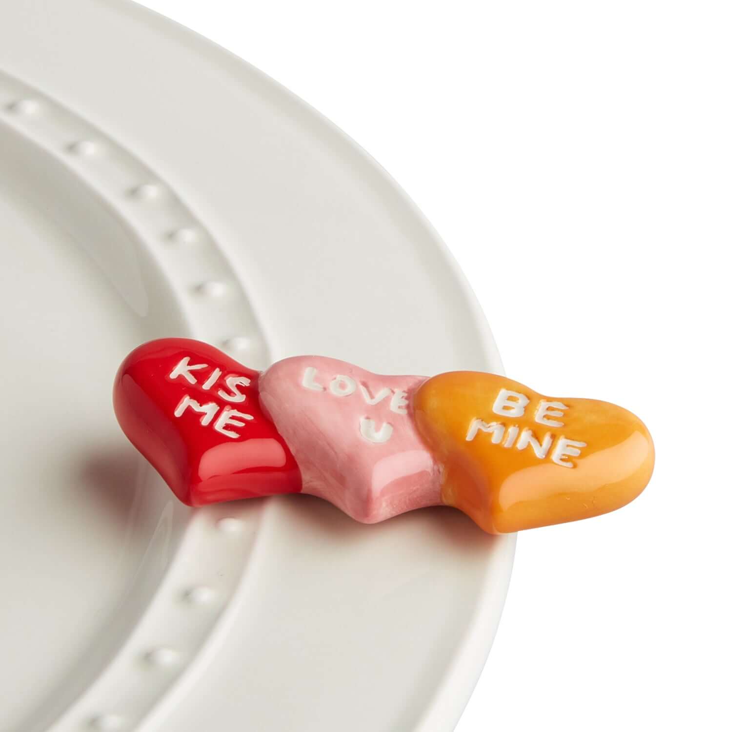 A set of three Nora Fleming Conversation Heart Mini A178 ceramic pieces with red and pink colors and the phrases 'KISS ME' and 'LOVE ME' inscribed on them, placed on a white plate.